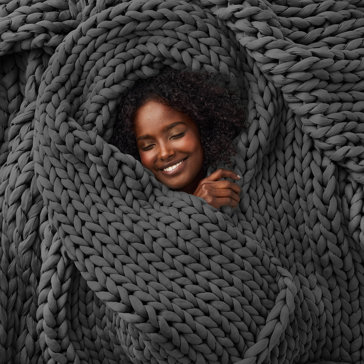 A woman peeking out from a large, knitted grey weighted blanket