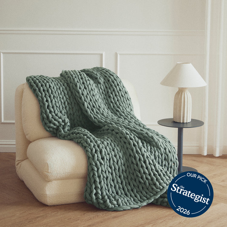 Botanical Dream bearaby chunky knit blanket draped over a beige armchair in a room with a lamp and side table.