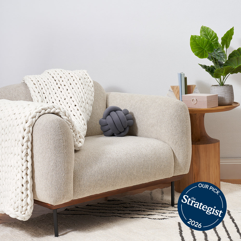 Cloud White knit blanket and gray knot pillow, next to a wooden side table with a plant and books.