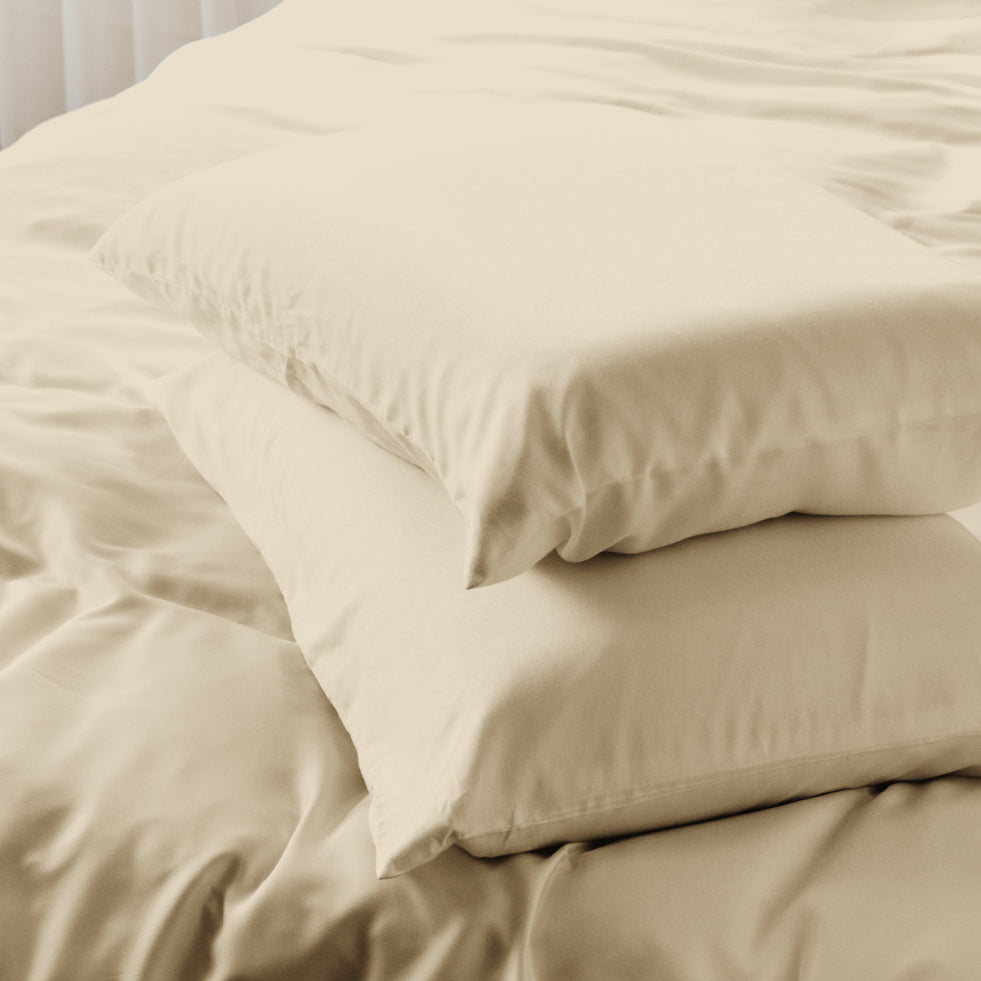 A stack of two bearaby driftwood pillows on a bed with matching cream bedding, shown in a softly lit bedroom setting.