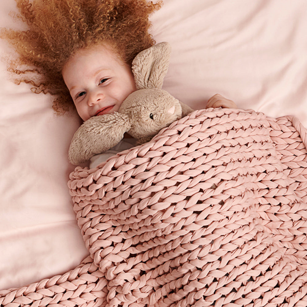 Child with curly hair holding a plush bunny under an kid evening rose knit weighted blanket