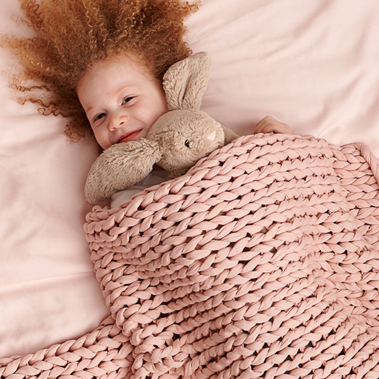 Child with curly hair holding a plush bunny under an kid evening rose knit weighted blanket