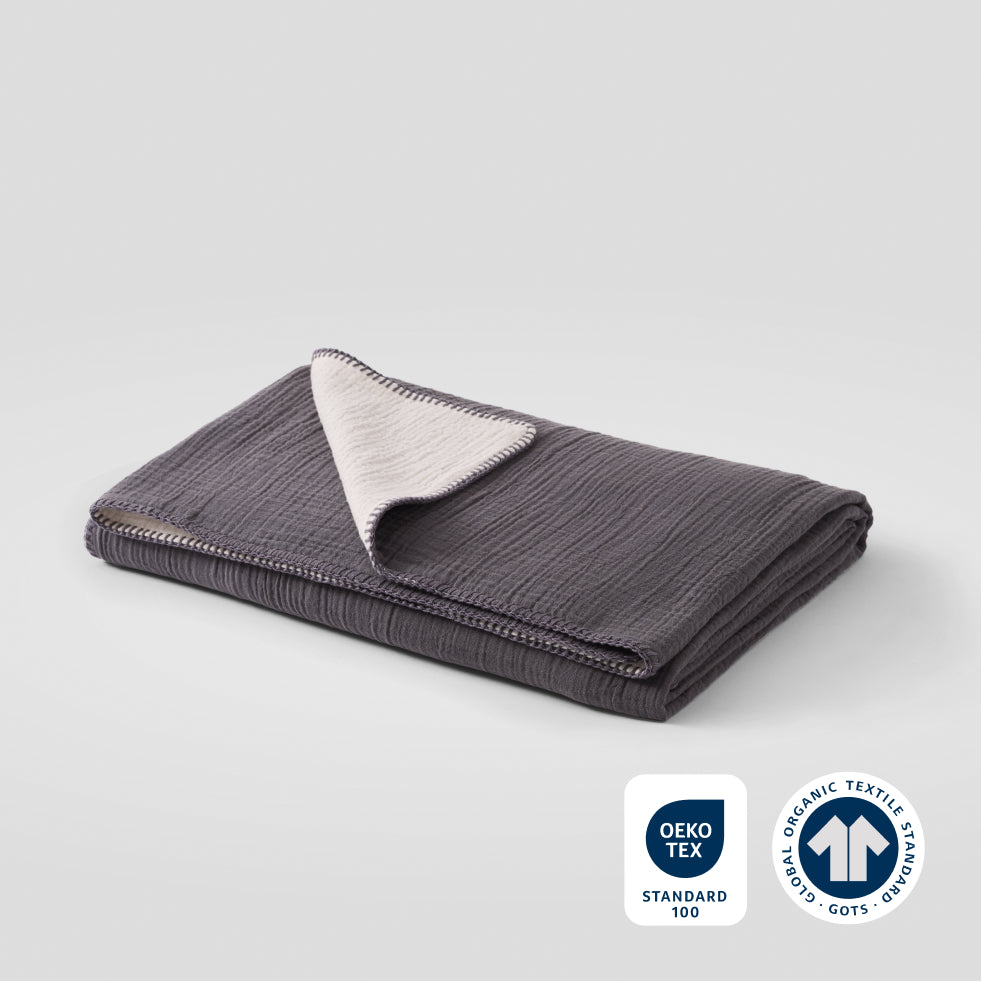 A folded asteroid color cotton muslin blanket with OEKO-TEX and GOTS certification badges in the lower right corner