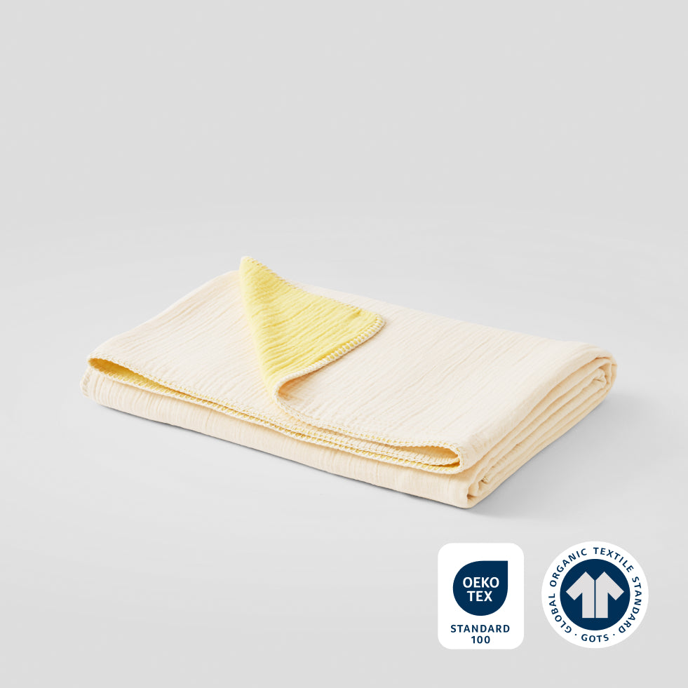 A folded lemon icing cotton muslin blanket with OEKO-TEX and GOTS certification badges in the lower right corner.
