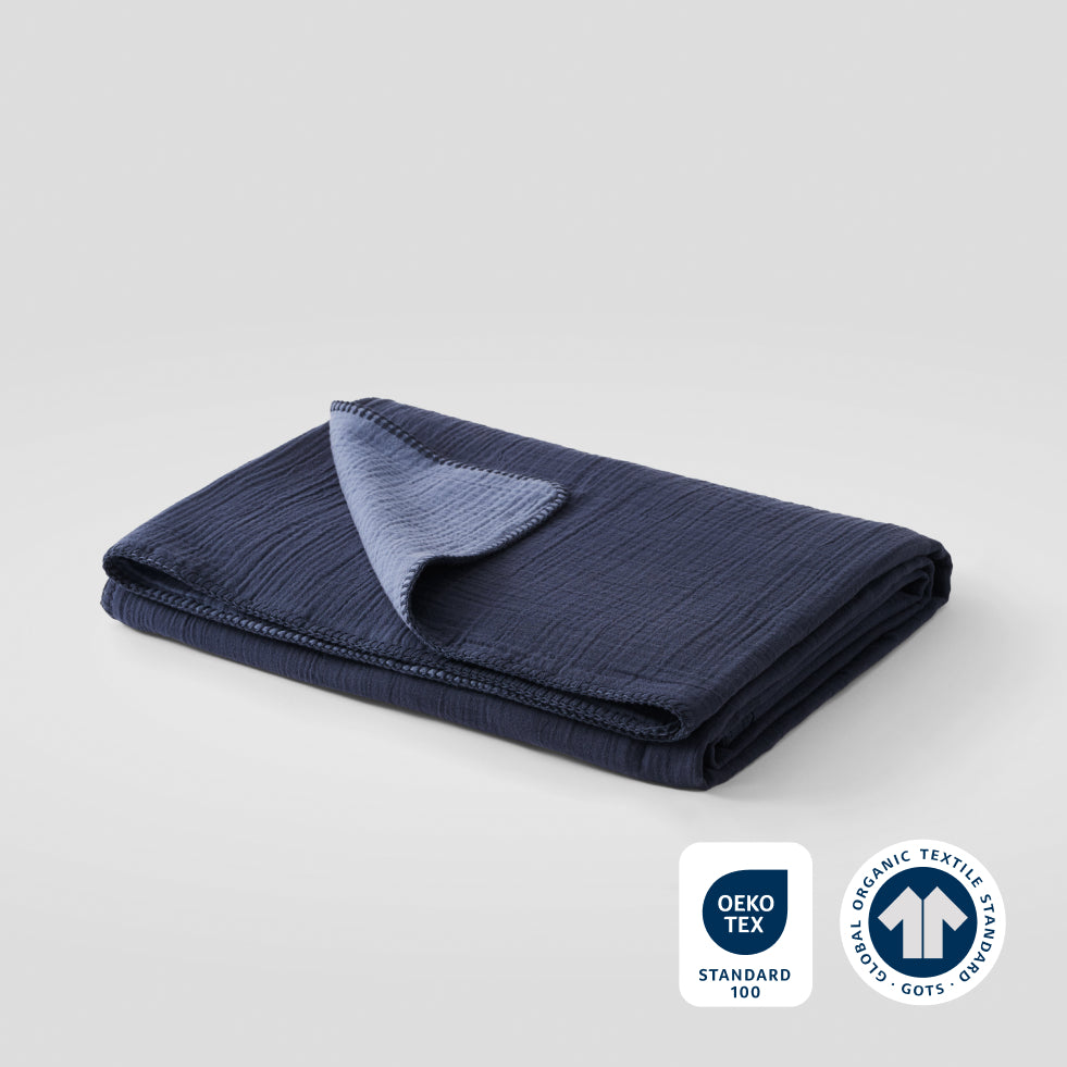 A folded midnight blue cotton muslin blanket with OEKO-TEX and GOTS certification badges in the lower right corner