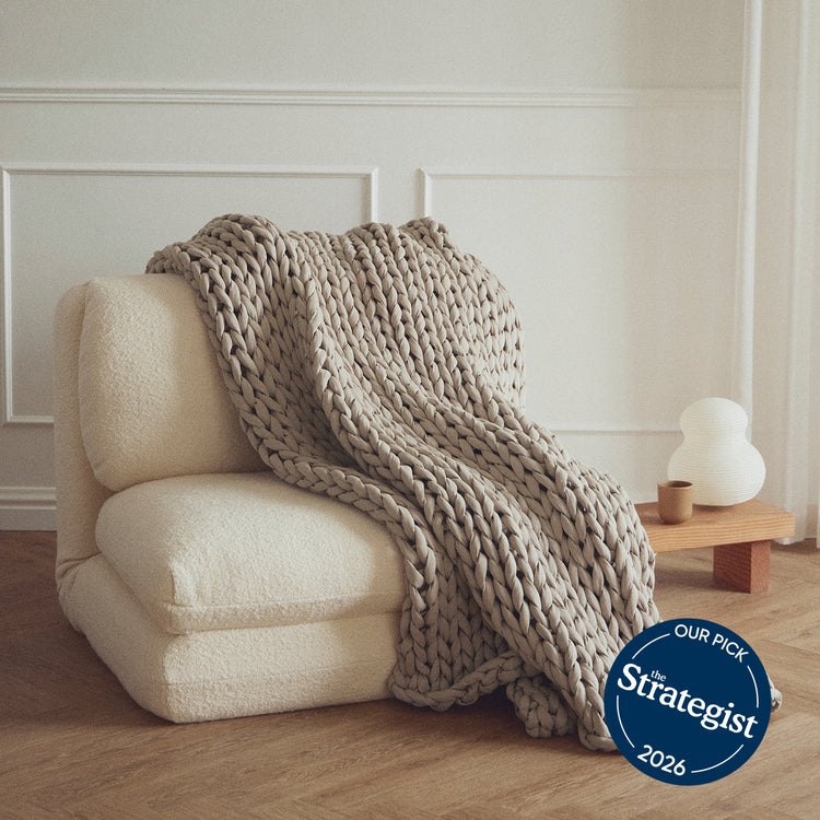Beige armchair with a bearaby chunky knit softwood blanket draped over it, in a room with a neutral color scheme.