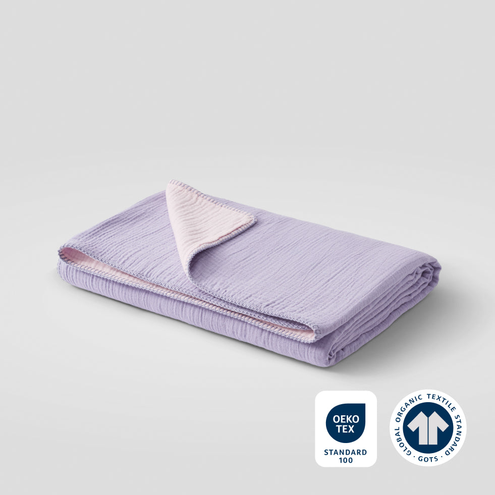 A folded sweet lavender cotton muslin blanket with OEKO-TEX and GOTS certification badges in the lower right corner